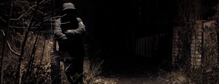 Mann mit dunkler Kapuzenmantel lehnt im Dunkeln an einer Metallstange.