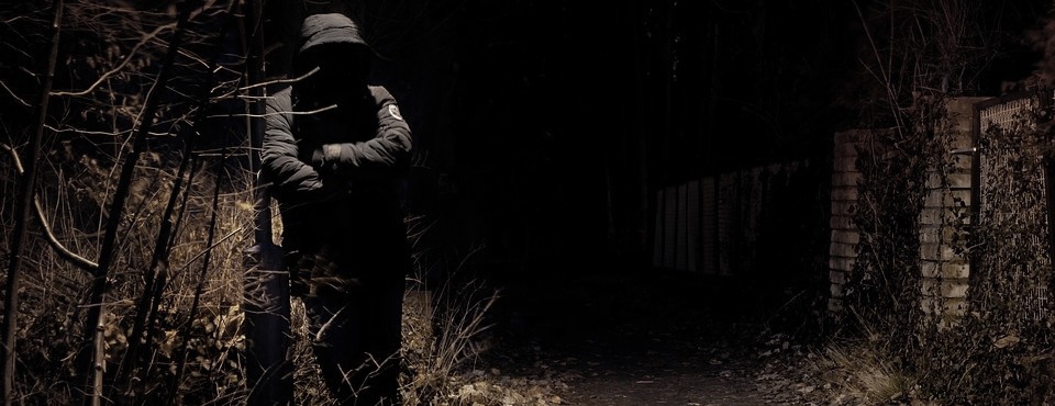 Mann mit dunkler Kapuzenmantel lehnt im Dunkeln an einer Metallstange.